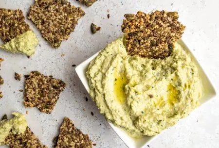 One small bowl of creamy artichoke dip, shown on bench top with broken seedy crackers for dipping