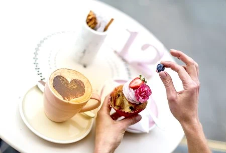 A cup and saucer with coffee and chocolate love heart dusted on top with a cupcake topped with pink icing, a pink flower and a strawberry and blueberry picked off by a hand in the picture