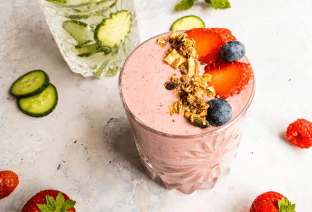 Two glass cups, one displaying berry smoothie with fresh fruit & the other with an elderflower cocktail mix with cucumber