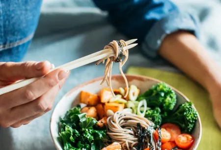 A bowl of noodles, veggies and prawns with someone wrapping the noodles around chopsticks in a denim shirt