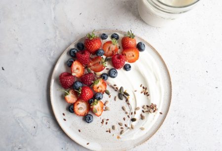 A glass pot of yogurt alongside two plates with yogurt, seeds and berries