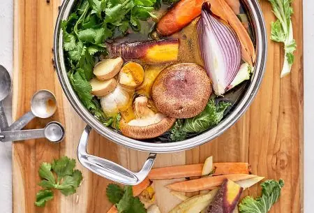 A wooden board with a pot of vegetables and broth and herbs inside