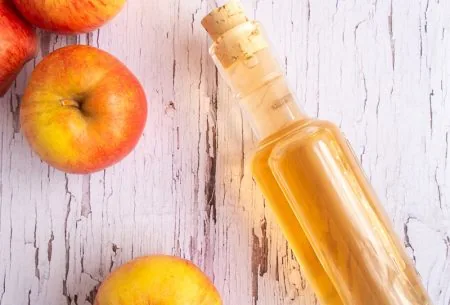 Apple cider vinegar in a glass bottle on white wooden surface with fresh apples alongside
