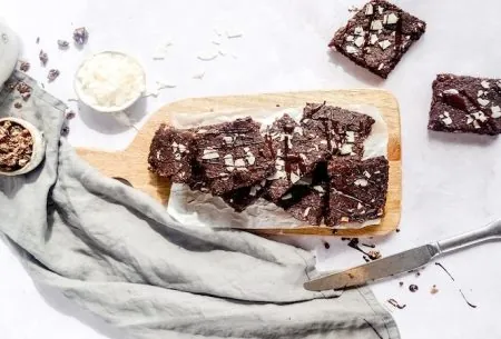 Eight chocolate and coconut brownies stacked along a chopping board, topped with coconut flakes