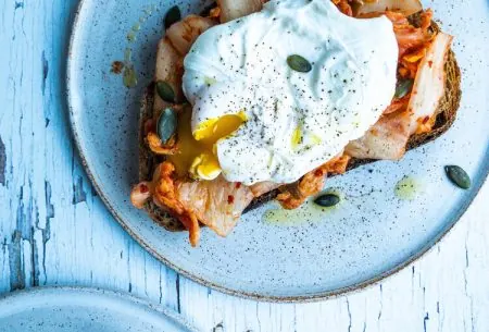 Two plates with one slice of toast kimchi and a poached egg with pumpkin seeds on top