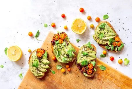 4 slices of toast topped with sliced avocado, fresh tomato and basil on a white surface and wooden chopping board with halved lemons and whole cherry tomatoes