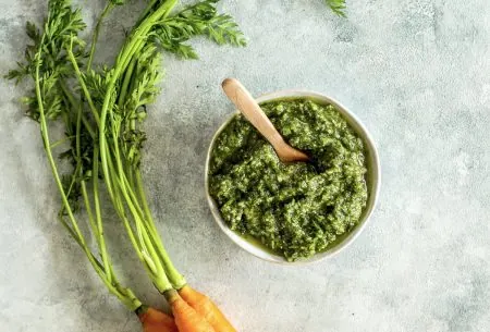 Pot of green carrot top pesto, shown on bench top with small bunch of fresh carrots and leafy green carrot tops