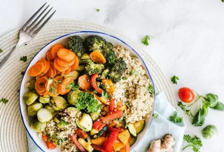 A colourful bowl of food including quinoa, roasted mixed peppers, courgette, carrot, brussell sprouts and broccoli on a place mat with a napkin and piece of ginger