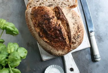 A loaf of sourdough that has been slices alongside a knife, salt, pepper, basil and a bottle of oil