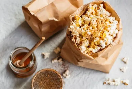 A brown paper bag of popcorn and a cinnamon shaker and a glass pot of honey with a honey spoon