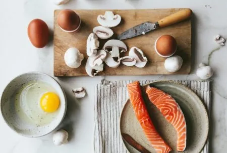 A wooden chopping board with sliced mushrooms, and whole eggs. A bowl with a cracked egg inside and a plate with two salmon fillets and a fish knife