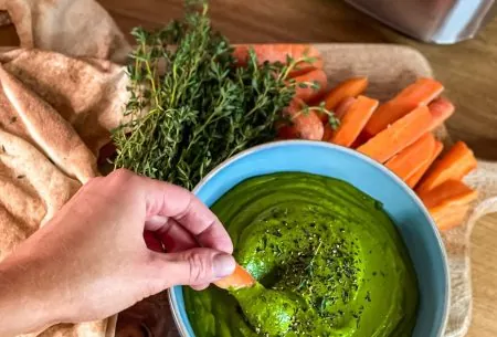 One small bowl full of pistachio and white bean dip. Carrot slices and fresh thyme accompanying, and a hand dipping into dip