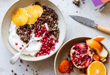 A bowl of yogurt topped with granola and sliced orange and pomegranate seeds and a bowl with the remaining half a pomegranate and orange