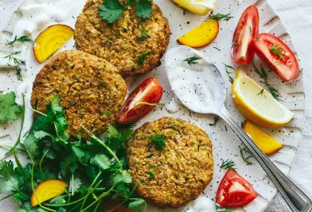 One plate with three cooked courgette and chickpea fritters, served with probiotic tzatziki, fresh greens and tomato