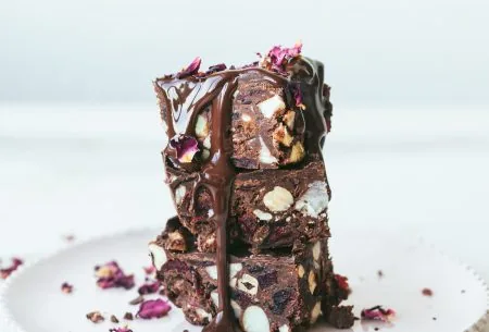 Three fruit and nut chocolate bites stacked on a white plate, drizzled with chocolate and garnished with small rose petals