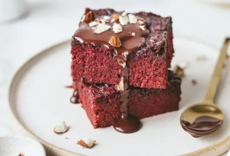 Two prebiotic beet velvet brownies stacked on a plate with melted chocolate and crushed almonds