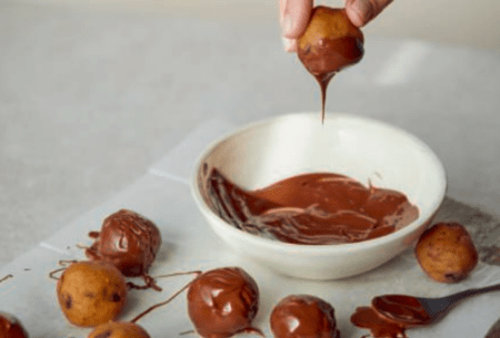 View The science of food cravings & what to do about them? A small bowl of melted chocolate with a hand dipping in a cookie dough ball to cover in chocolate, other balls already covered in chocolate and not covered in chocolate are alongside in rows