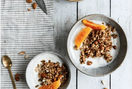 Two bowls of yogurt topped with granola and blood orange on a white wooden surface with a black and white striped tea towel, a spoon and a knife and a plate with segments of orange