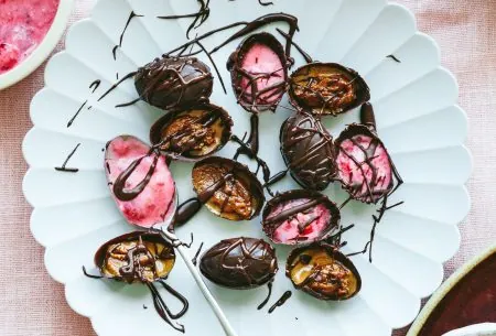 Six small chocolate almond Easter eggs on a white plate, cracked in half to show fillings and chocolate drizzle
