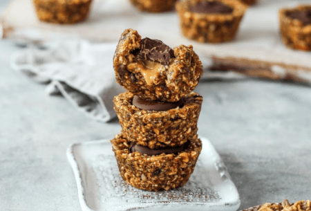 Three dark chocolate and peanut butter granola cups stacked on a small plate, with seven more scattered on bench top in background