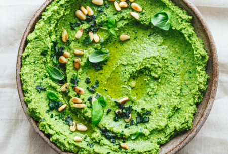 Bowl of bright green leafy pesto hummus, sprinkled with pine nuts and fresh basil leaves