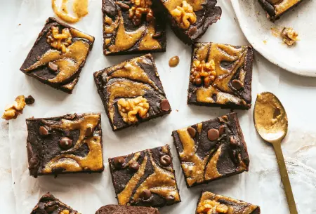 12 fudgy black bean and walnut brownies scattered on a bench top, drizzled with walnut butter