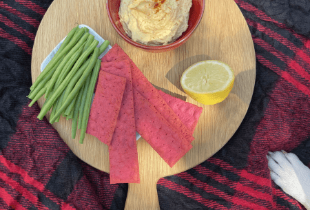 Circular chopping board on picnic rug with a small bowl of white bean hummus, crackers, green beans and lemon