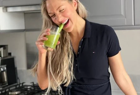 Dr Megan Rossi standing in a kitchen holding a celery juice up to her mouth and laughing