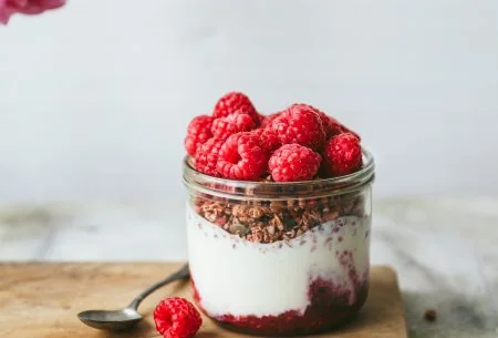 One yogurt pot with creamy live yogurt and crunchy granola, topped with fresh raspberries and displayed on a chopping board