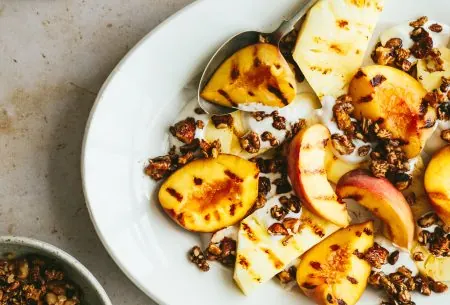 White plate with grilled peaches and walnut crumble, with an extra pot of walnuts to the side