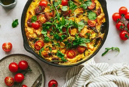 Cast iron skillet with cooked Mediterranean frittata and sundried tomatoes, shown with fresh cherry tomatoes on bench top