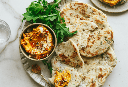 Two fresh 'live' and herby flatbreads on a plate with a pot of curried sweet potato dip and fresh salad greens