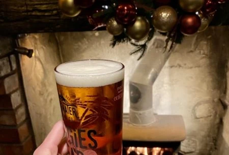 One hand holding up a glass full with beer, festive fireplace and decorations shown in background