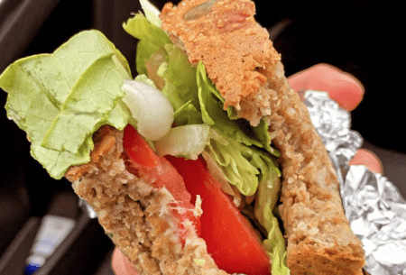Close up image of a hand holding a homemade sandwich with visible lettuce and tomato, wrapped in foil