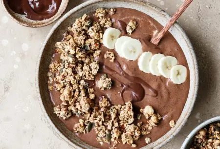 Chocolate and banana smoothie bowl topped with extra granola and banana, and a small pot of extra chocolate