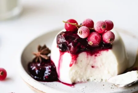 One serve of live panna cotta with mulled berry compote on top and frozen berries, served on a small ceramic plate