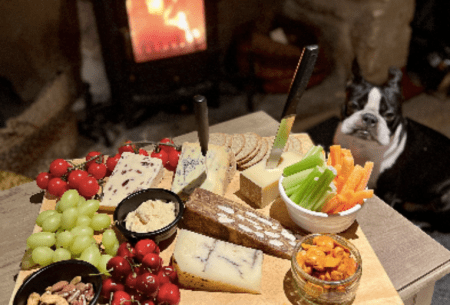 Platter of assorted food including fruits and cheeses set out on chopping board with fireplace in background
