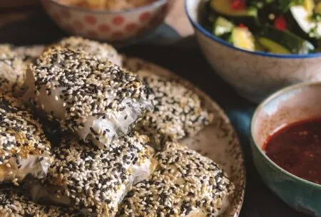 Rice paper dumplings covered in black and white sesame seeds on a plate with smaller plates in the background containing dips and salad