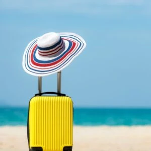 Holiday photo with suitcase and sun hat on a white beach with blue sky 