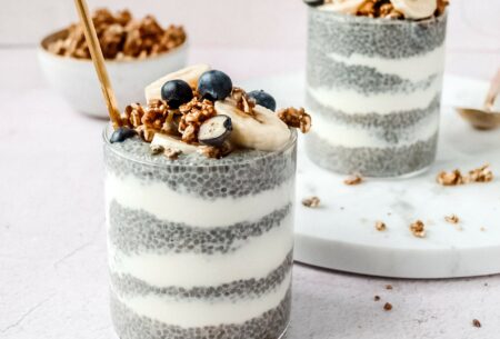 A picture of two clear glass tumblers with thick layers of plain yoghurt and chia seeds with a sprinkle of granola, sliced bananas and blueberries on top