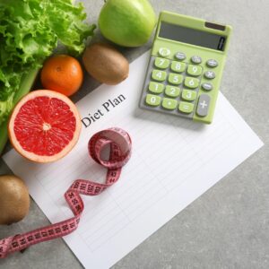 Photo of a green calculator, measuring table and some vegetables and fruit on a grey top table to reflect calorie counting
