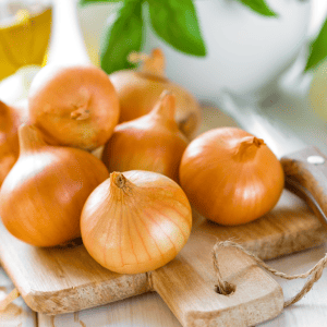 Photo of a bunch of brown onions on a wooden block with a plant in the background