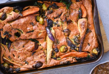A baking tray on a table with the vegetable toad-in-the-hole recipe inside. It has lots of vegetables like carrots, parsnips and olives on it as well as a relish on the side.