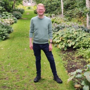 A photo of a man standing in a garden, he's wearing glasses and smiling