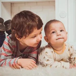 A photo of a baby and child side by side, lying down on the floor