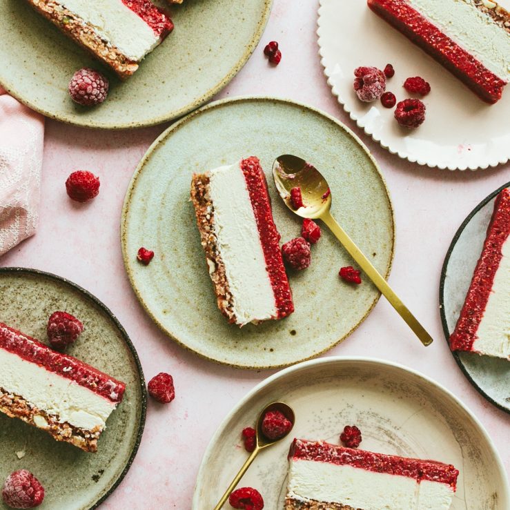 Three slices of raspberry and cashew cheesecake on light green plates, with fresh raspberries scattered