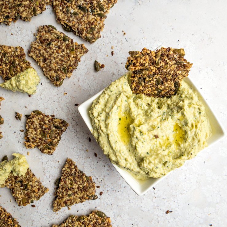 One small bowl of creamy artichoke dip, shown on bench top with broken seedy crackers for dipping