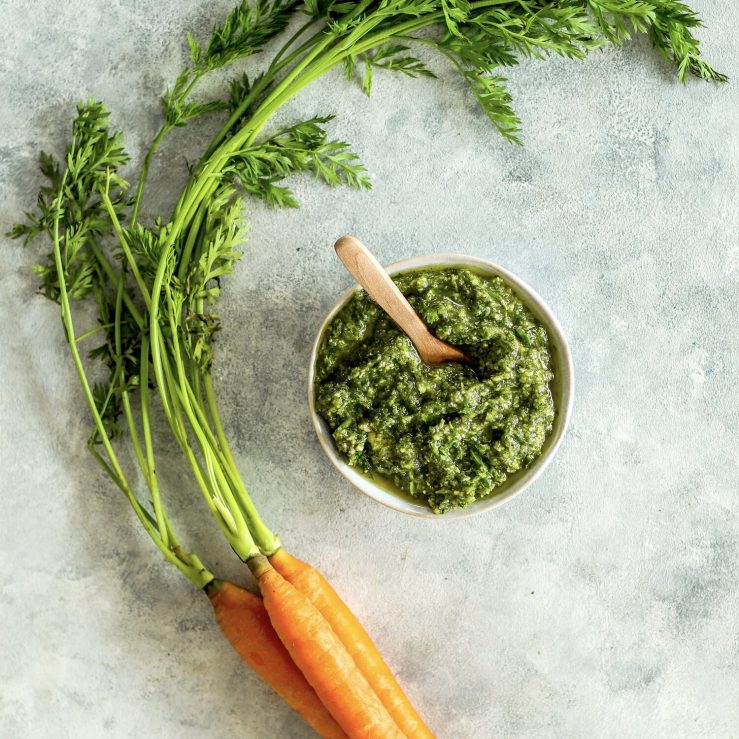 Pot of green carrot top pesto, shown on bench top with small bunch of fresh carrots and leafy green carrot tops