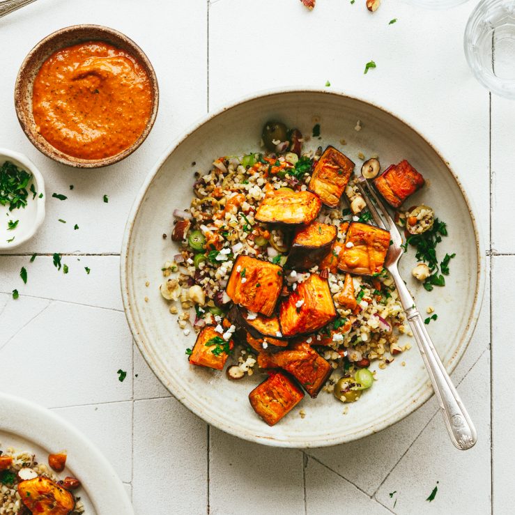 Roast aubergine with mixed grains and red pepper sauce, on a white place with a pot of extra sauce