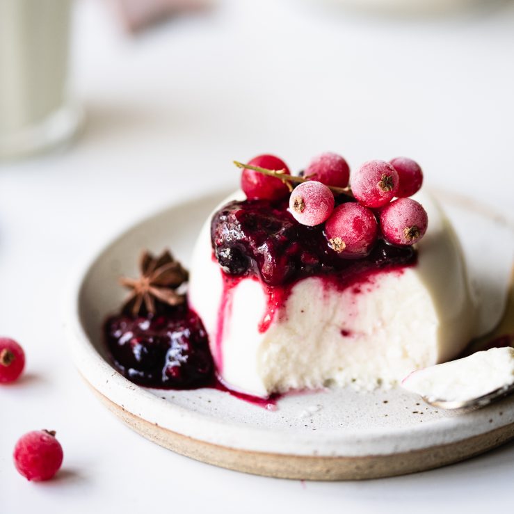 One serve of live panna cotta with mulled berry compote on top and frozen berries, served on a small ceramic plate